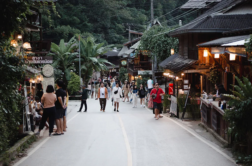 Dusk street scene with glowing lanterns and visitors walking between wooden shops in Mae Kampong