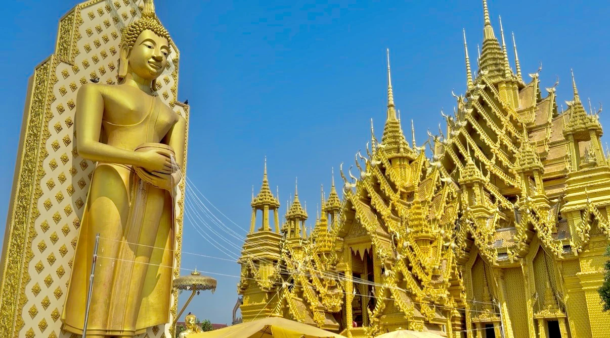 Standing golden Buddha with alms bowl beside the gilded viharn at Wat Chan Tawan Tok