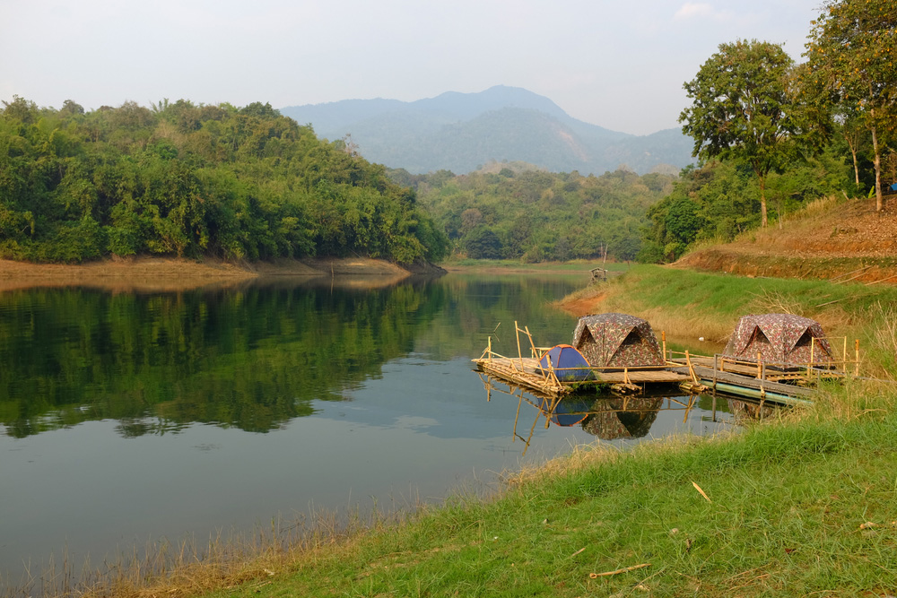 Floating tents on bamboo rafts beside the still waters of the Vajiralongkorn Reservoir, with forested mountains beyond