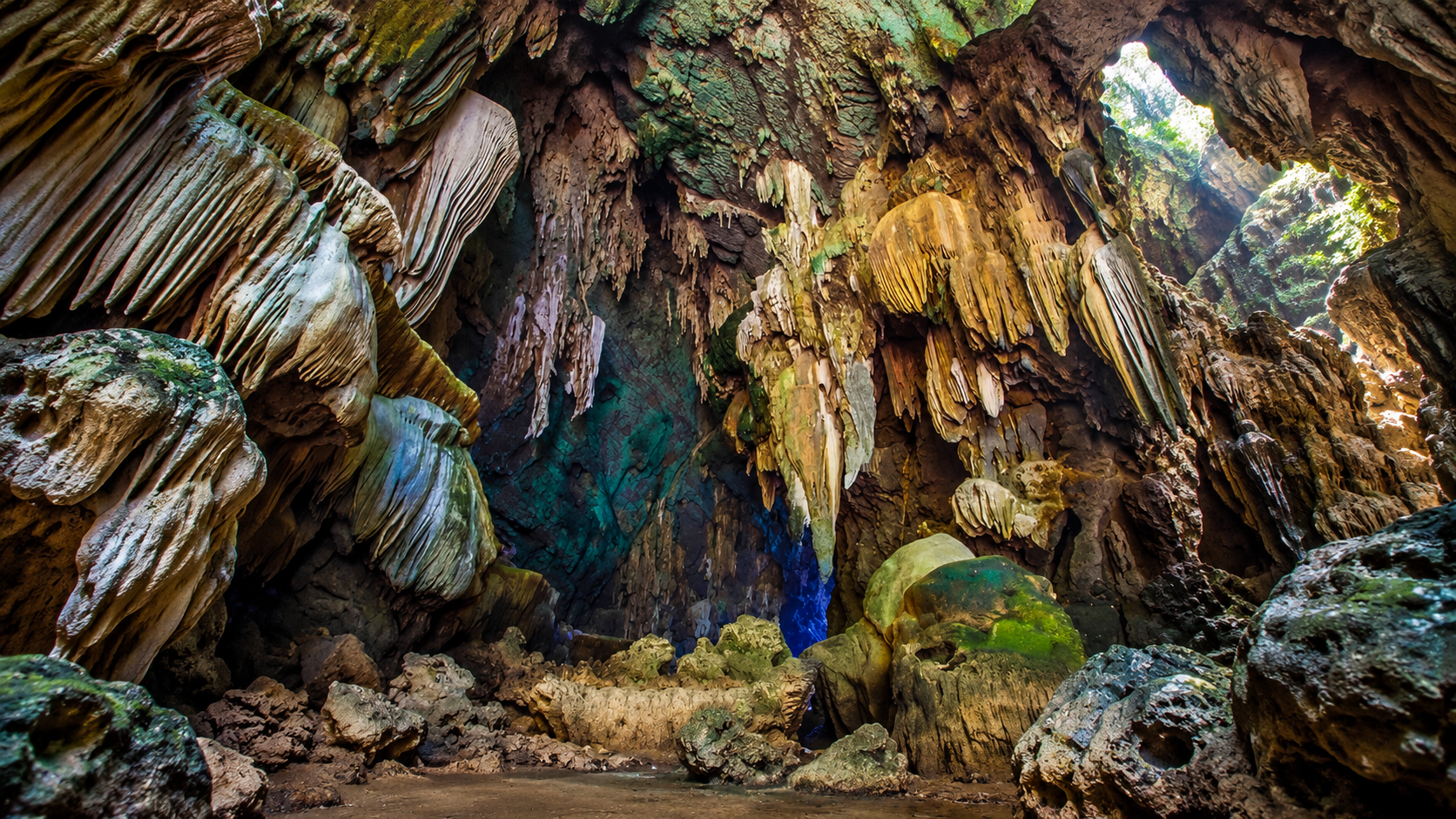A wide cave chamber of flowstone draperies, with daylight entering through an opening in the roof at Lam Khlong Ngu National Park