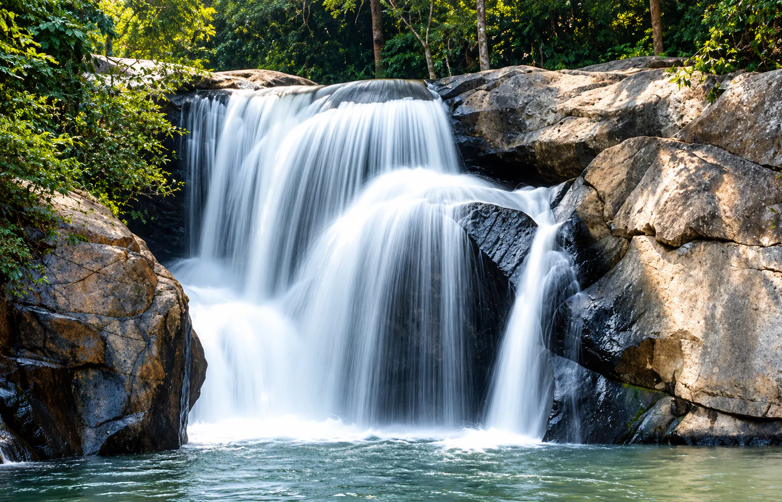A forest waterfall spilling over dark rock into a green pool, in the wooded landscape around Si Satchanalai, Sukhothai