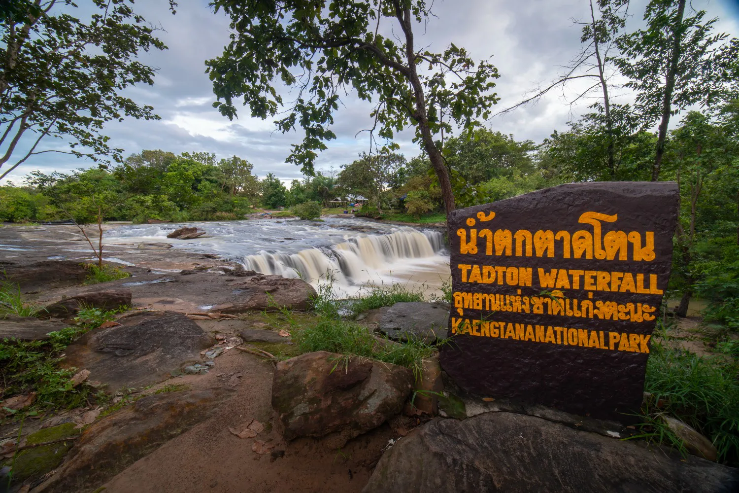 Water moving across a broad rock shelf into a shallow cascade, with riverside trees and a stone waterfall sign in the foreground