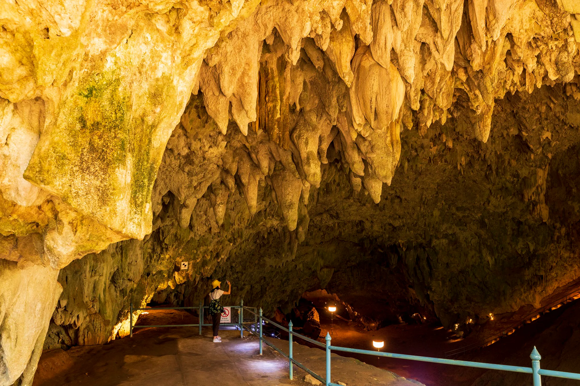 A visitor photographing dense stalactites along a railed walkway inside a limestone cave at Tham Luang–Khun Nam Nang Non, Chiang Rai