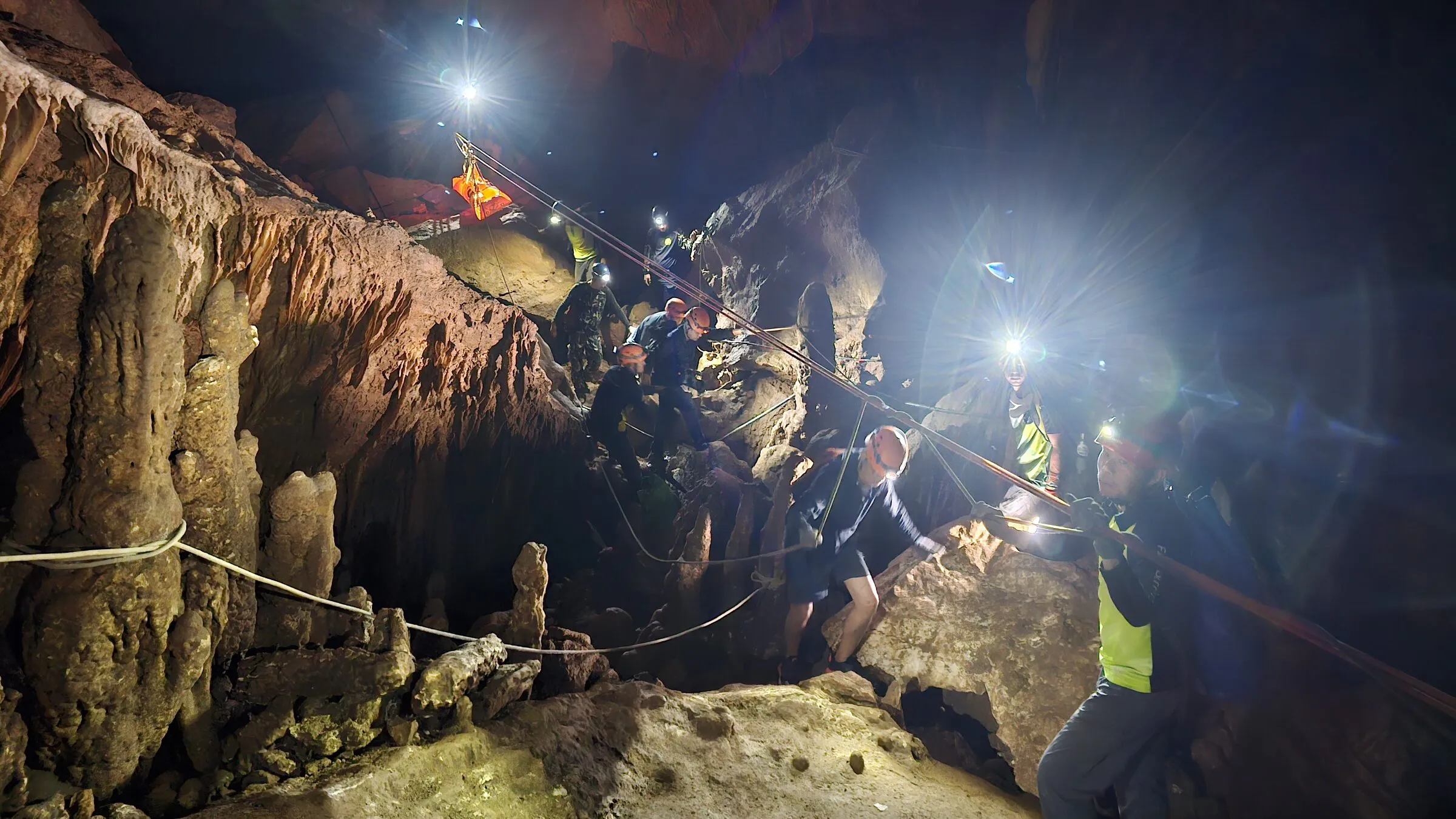 People with headlamps and ropes moving through a narrow, brightly lit limestone passage inside Tham Luang Cave