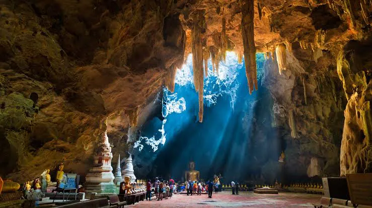 Shaft of daylight through a high cave ceiling onto a golden Buddha and white chedis among stalactites at Tham Luang–Khun Nam Nang Non, Chiang Rai