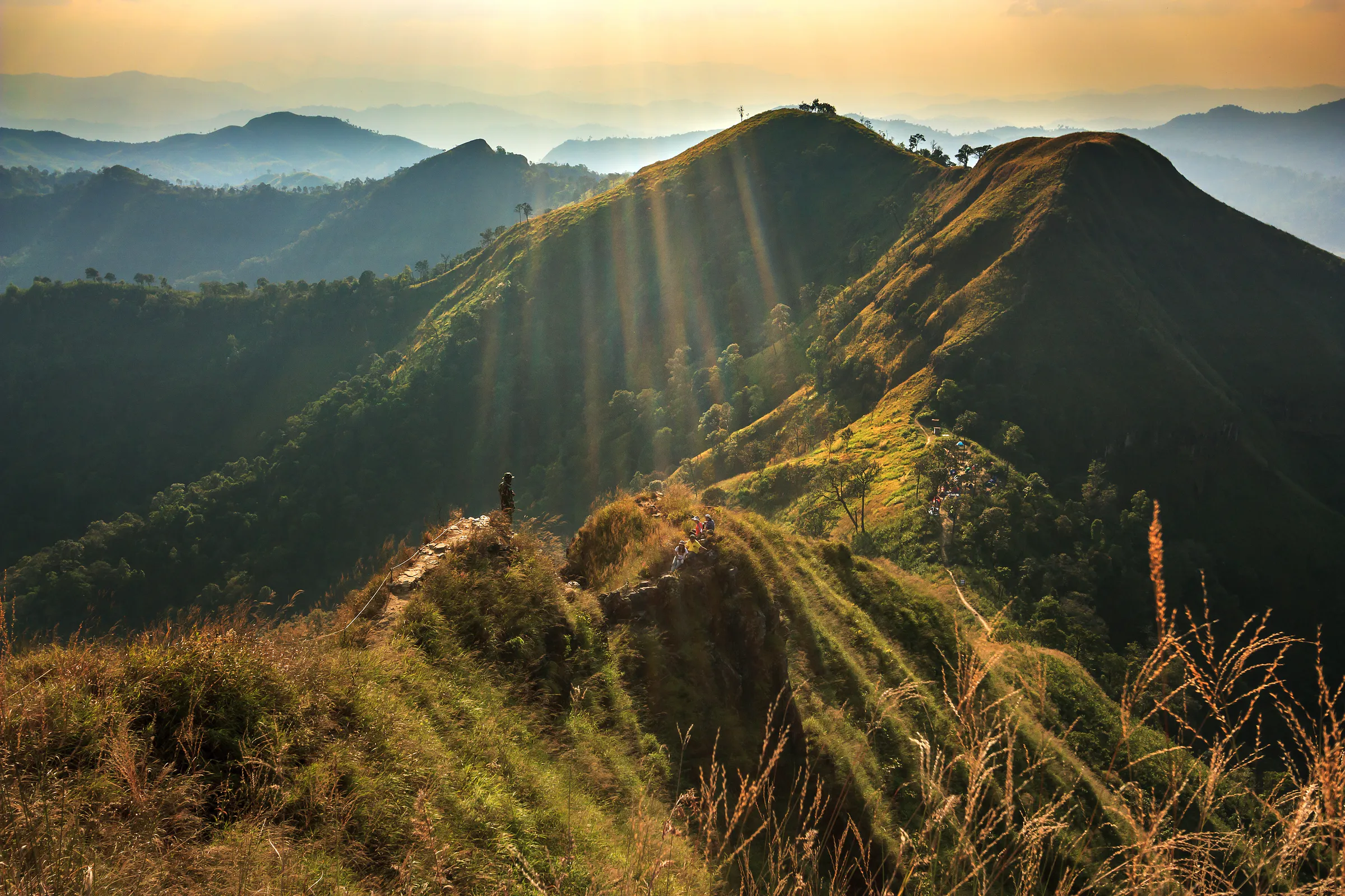 Hikers on the grassy ridge of Khao Chang Phueak in Thong Pha Phum National Park, Kanchanaburi, with sunbeams over layered western mountains