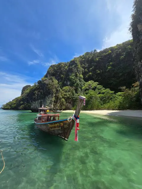 Traditional long-tail boat on clear emerald water beside a forested limestone cliff